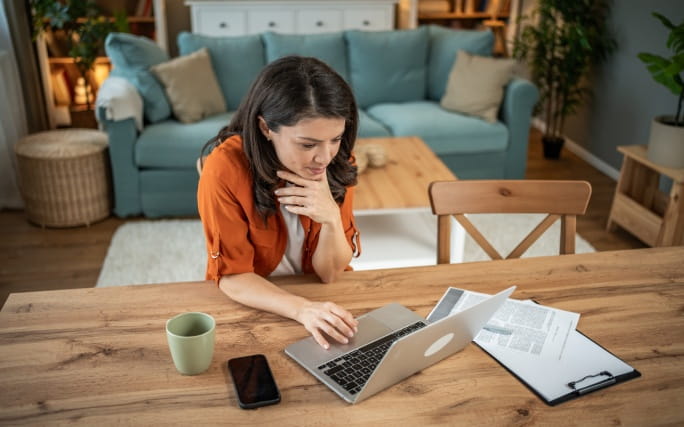 Mulher na sala de casa, sentada à mesa usa um computador portátil.